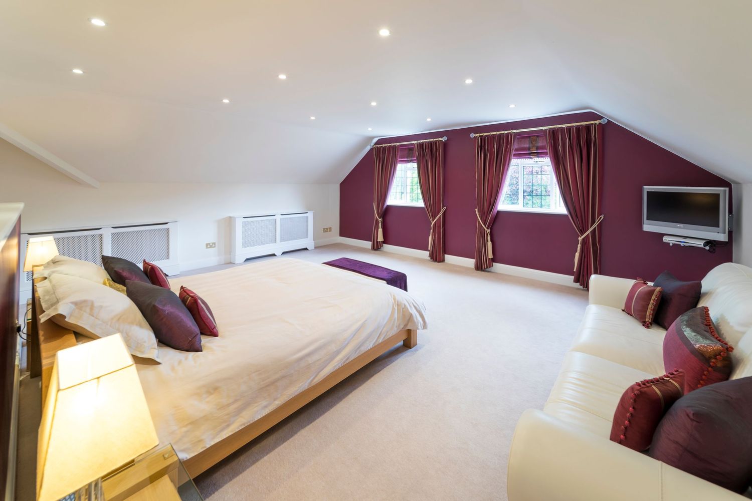 Spacious loft bedroom with burgundy accent walls, cream carpet, large bed, curtained skylights, and a wall-mounted TV.