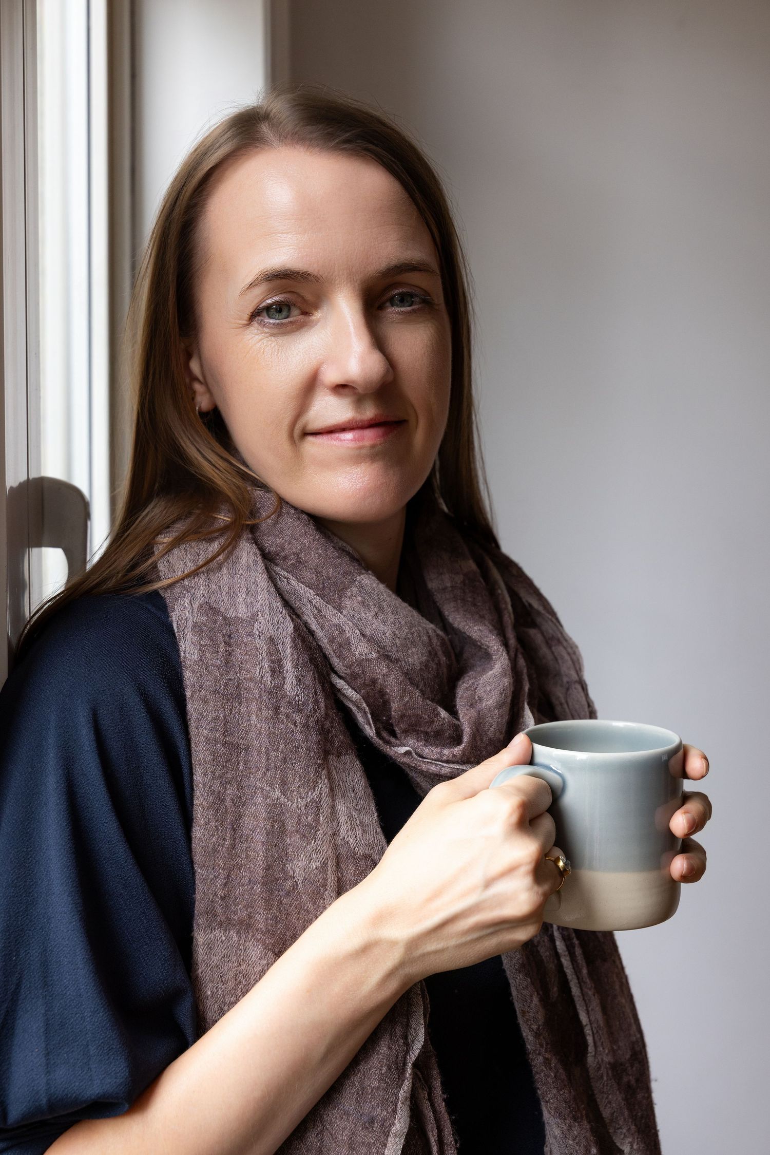 Woman holding a gray ceramic mug near a window, wearing a navy top and brown scarf, smiling softly.