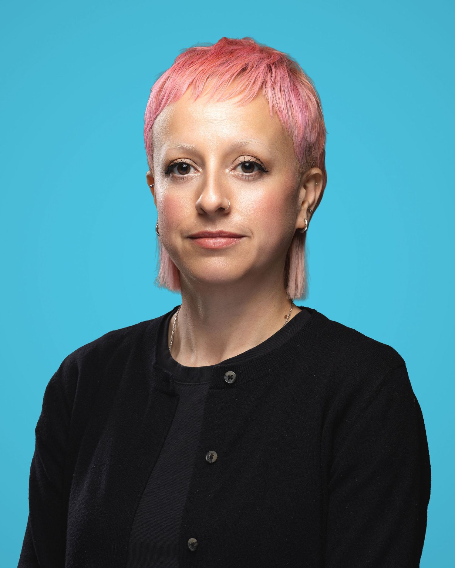 Woman with short pink hair wearing a black cardigan poses against a bright blue background.