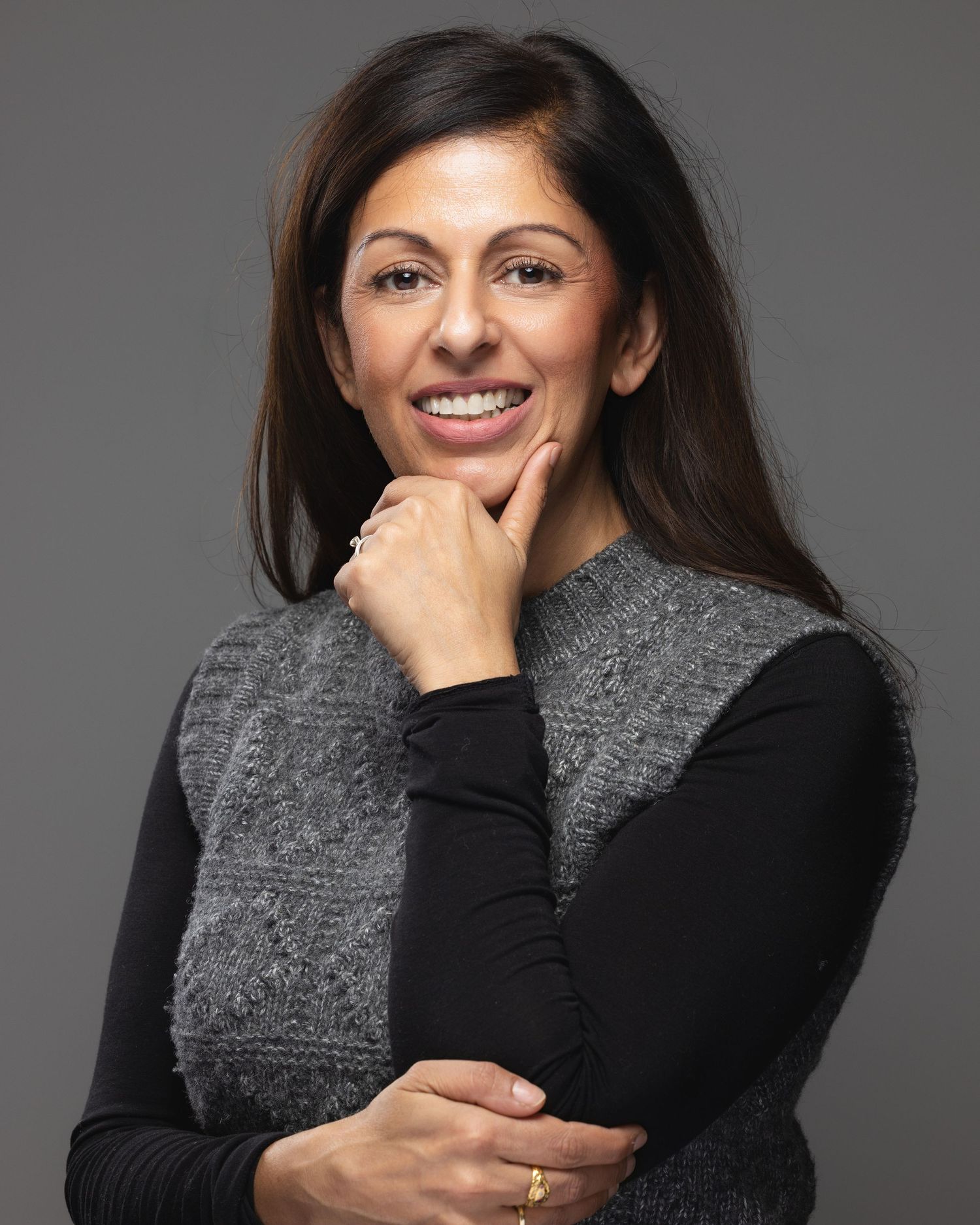 Professional woman with dark hair smiling confidently, wearing a gray vest over black top, posing against gray background.