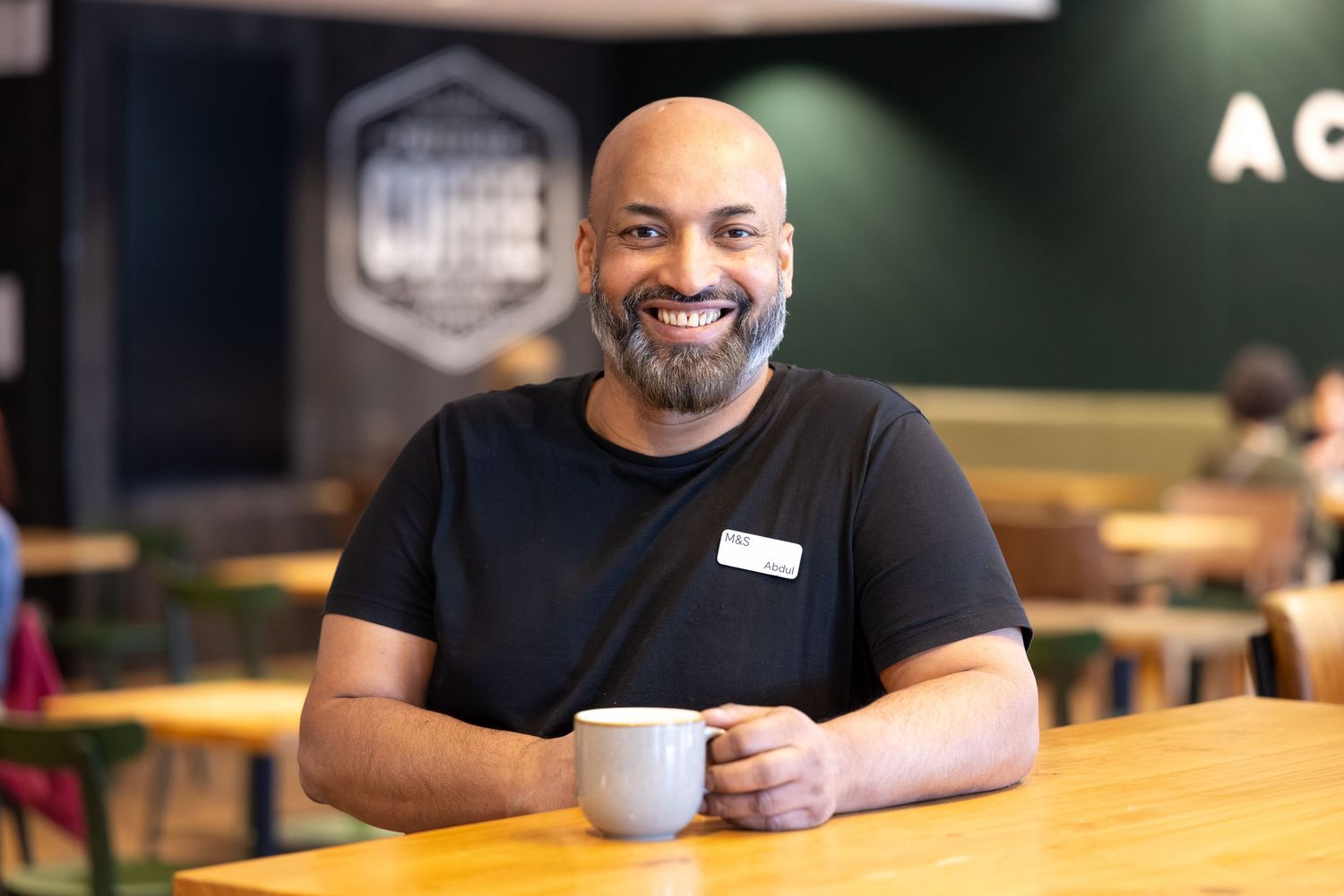 Corporate lifestyle photography of a smiling professional holding a coffee mug at a wooden table in a modern café.