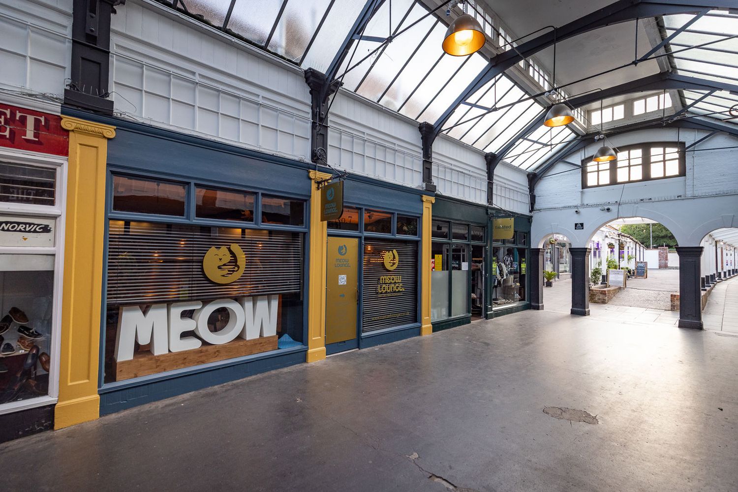 Shopping arcade interior with 'MEOW' signage on a storefront beneath a glass ceiling with natural lighting.