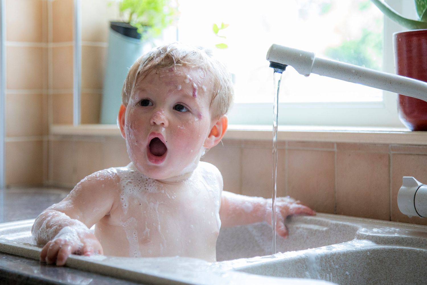 Cute toddler boy with wet hair taking a bubble bath in a kitchen sink with running water.