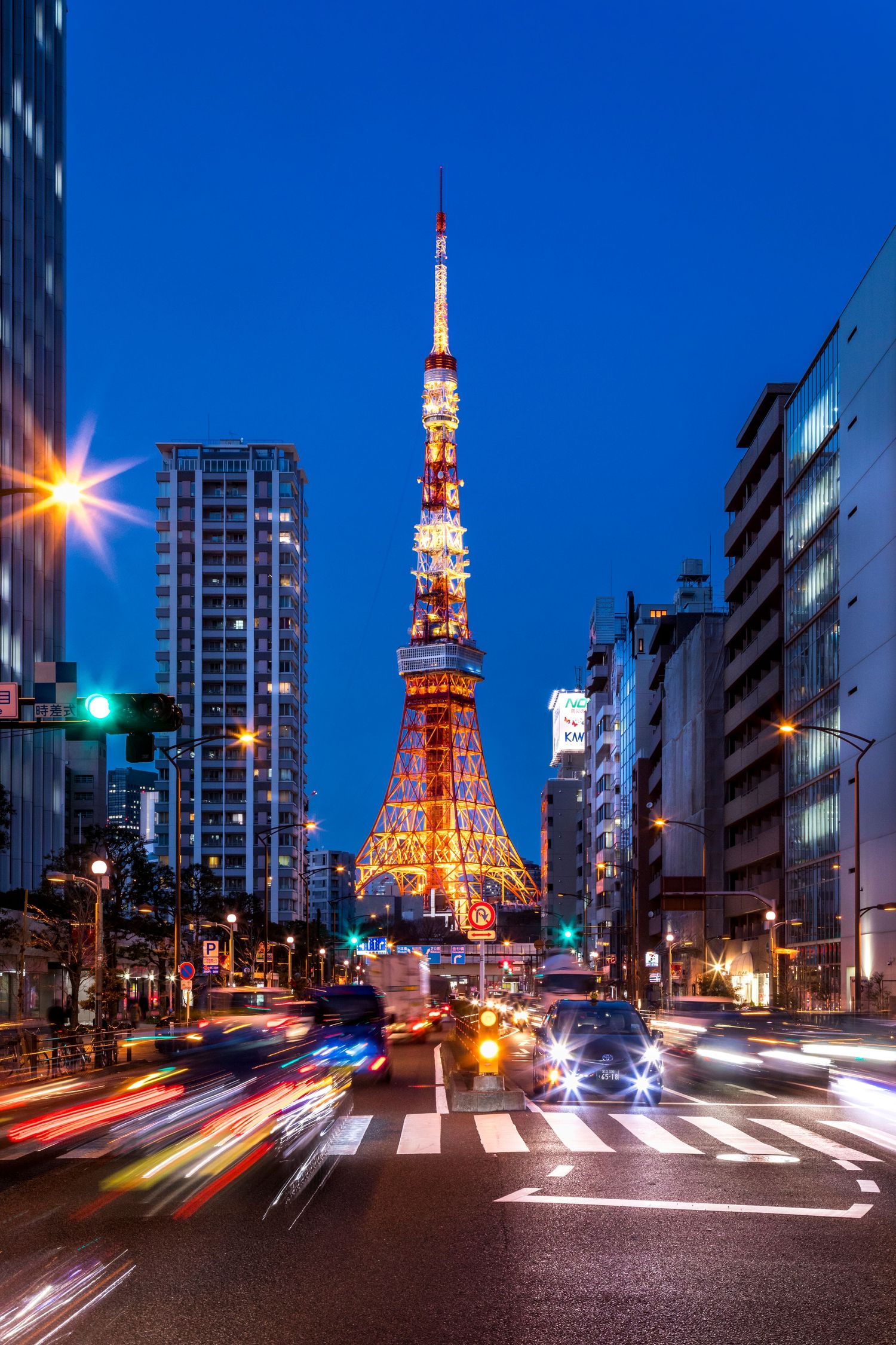 Tokyo Tower illuminated at night with light trails from traffic on busy urban street below.