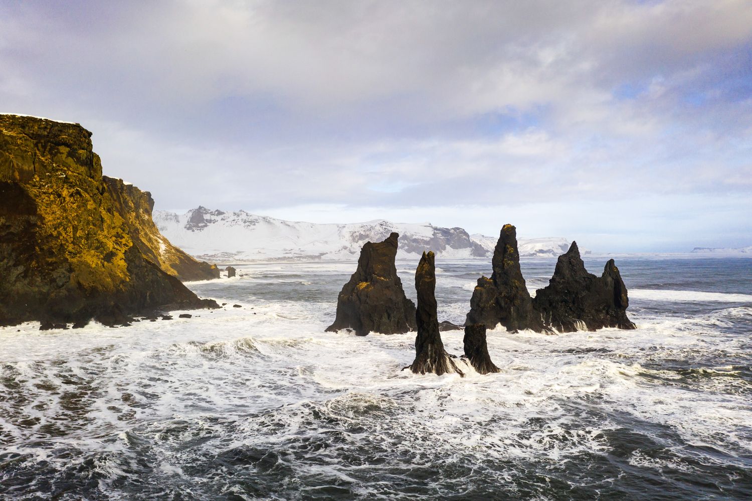 Dramatic sea stacks rise from turbulent waves at Vik black sand beach in Iceland under cloudy winter skies.