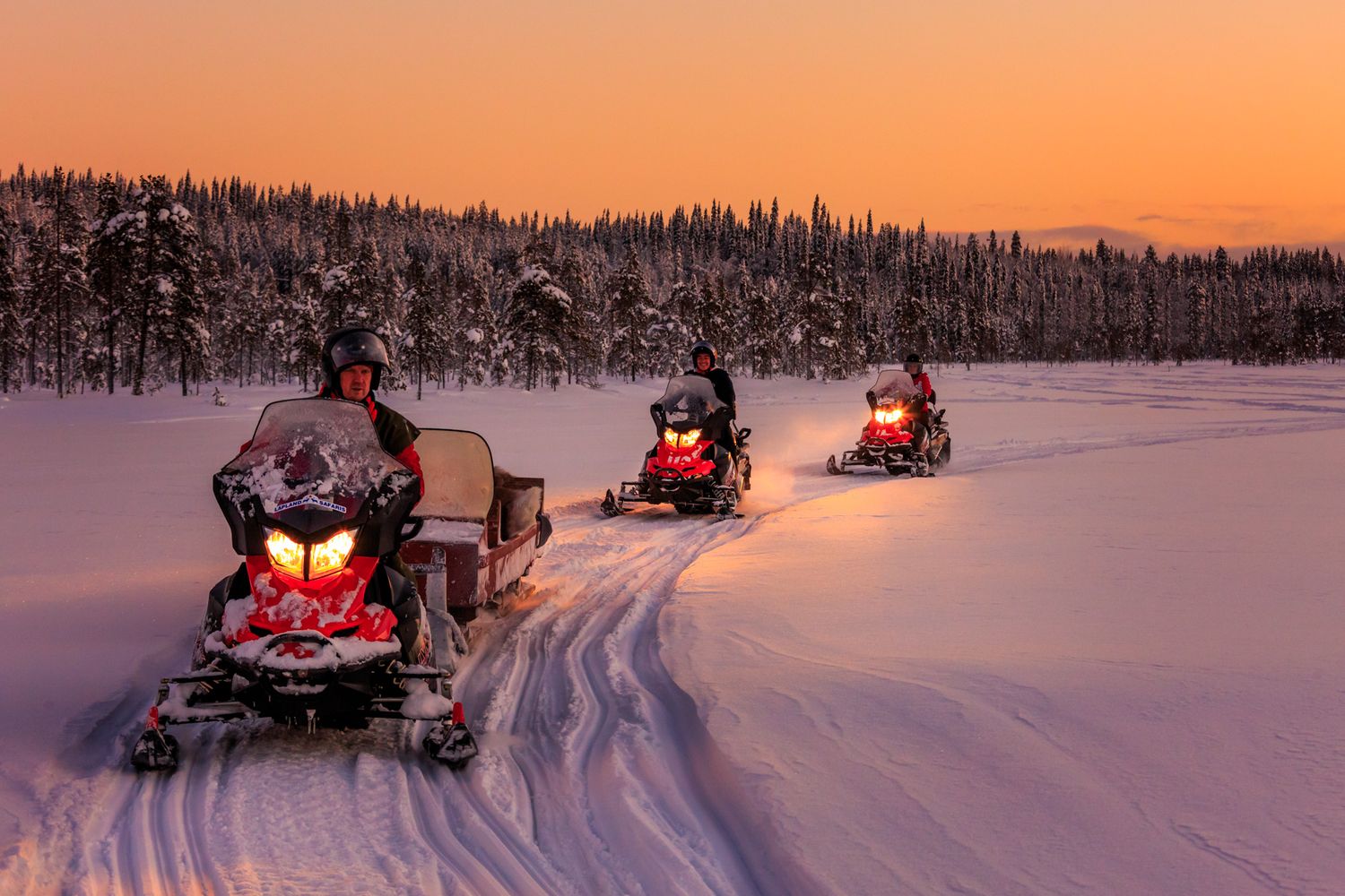 Snowmobilers ride across a snowy landscape at sunset with orange sky and snow-covered forest in background.