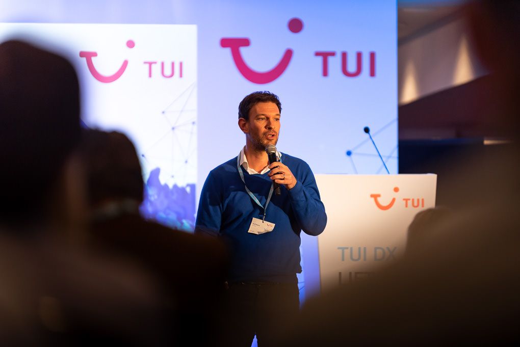 A presenter speaks on stage at a TUI corporate event with pink and blue branded backdrop lighting. Shot at a conference in London.