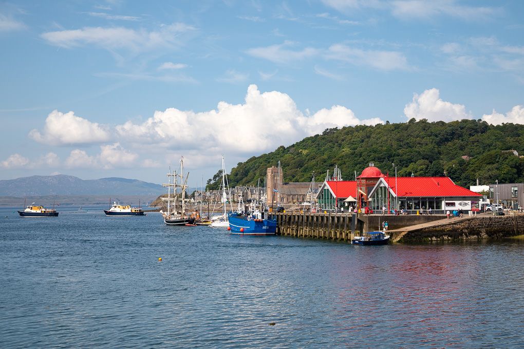 Scenic harbour view of Oban, Scotland, with fishing boats docked near red-roofed buildings along the waterfront.
