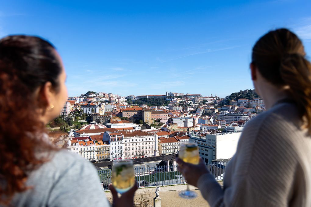 Two people look out over the historic cityscape of Lisbon, Portugal with white buildings under bright blue skies.