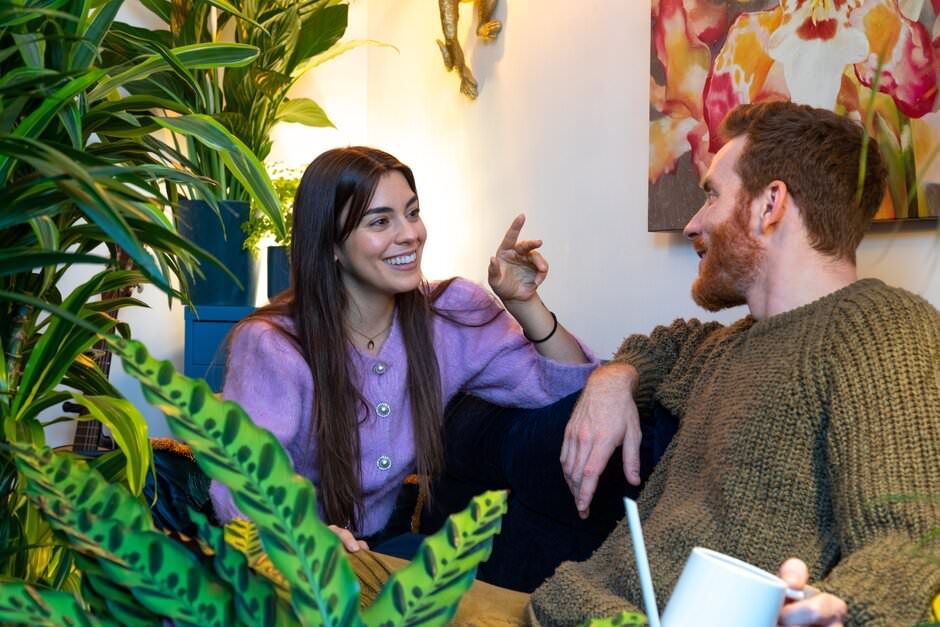 A lifestyle shoot of a young couple sitting close together on a couch surrounded by lush green houseplants.