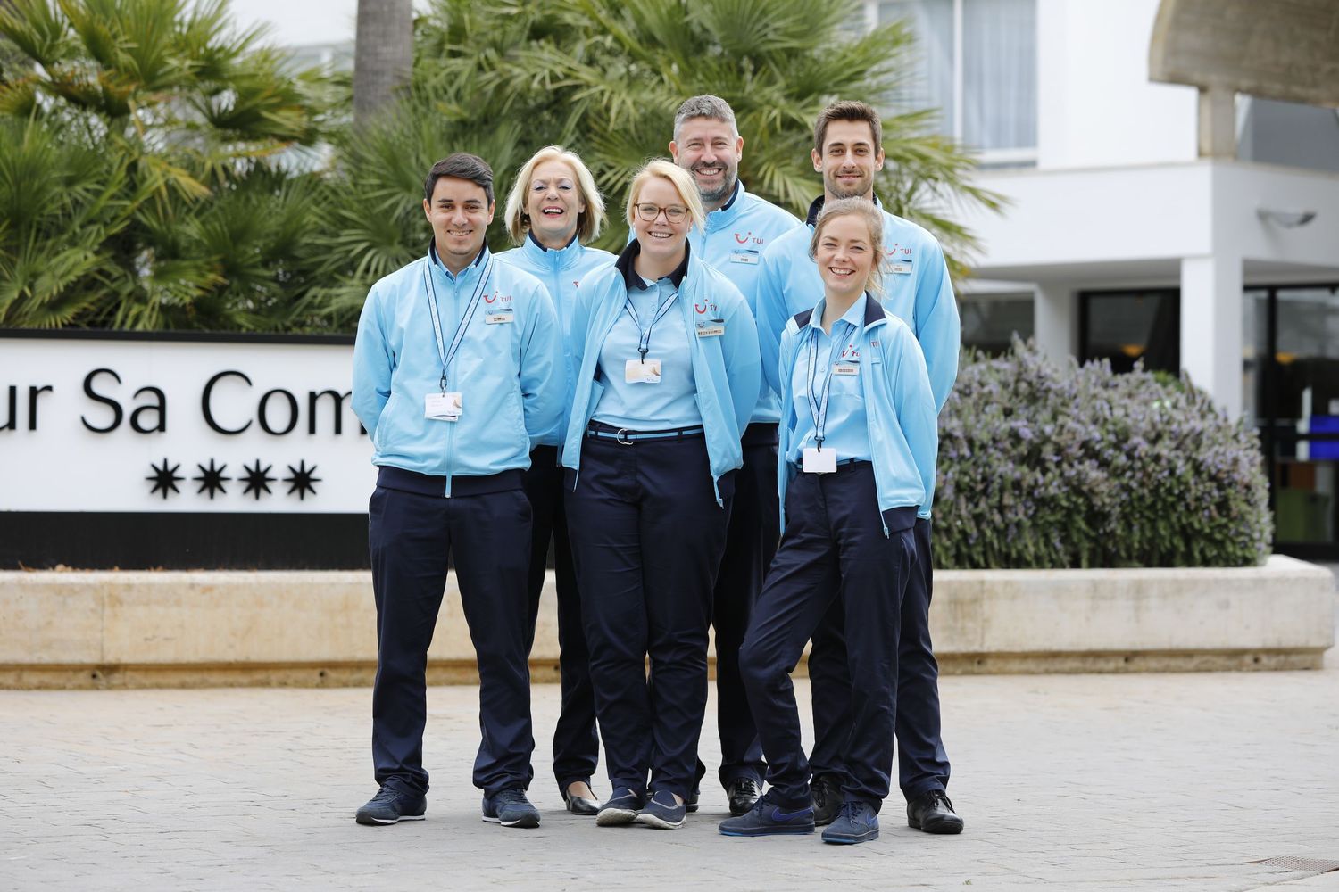 Six hotel staff in light blue uniforms smiling together outside a four-star resort entrance.