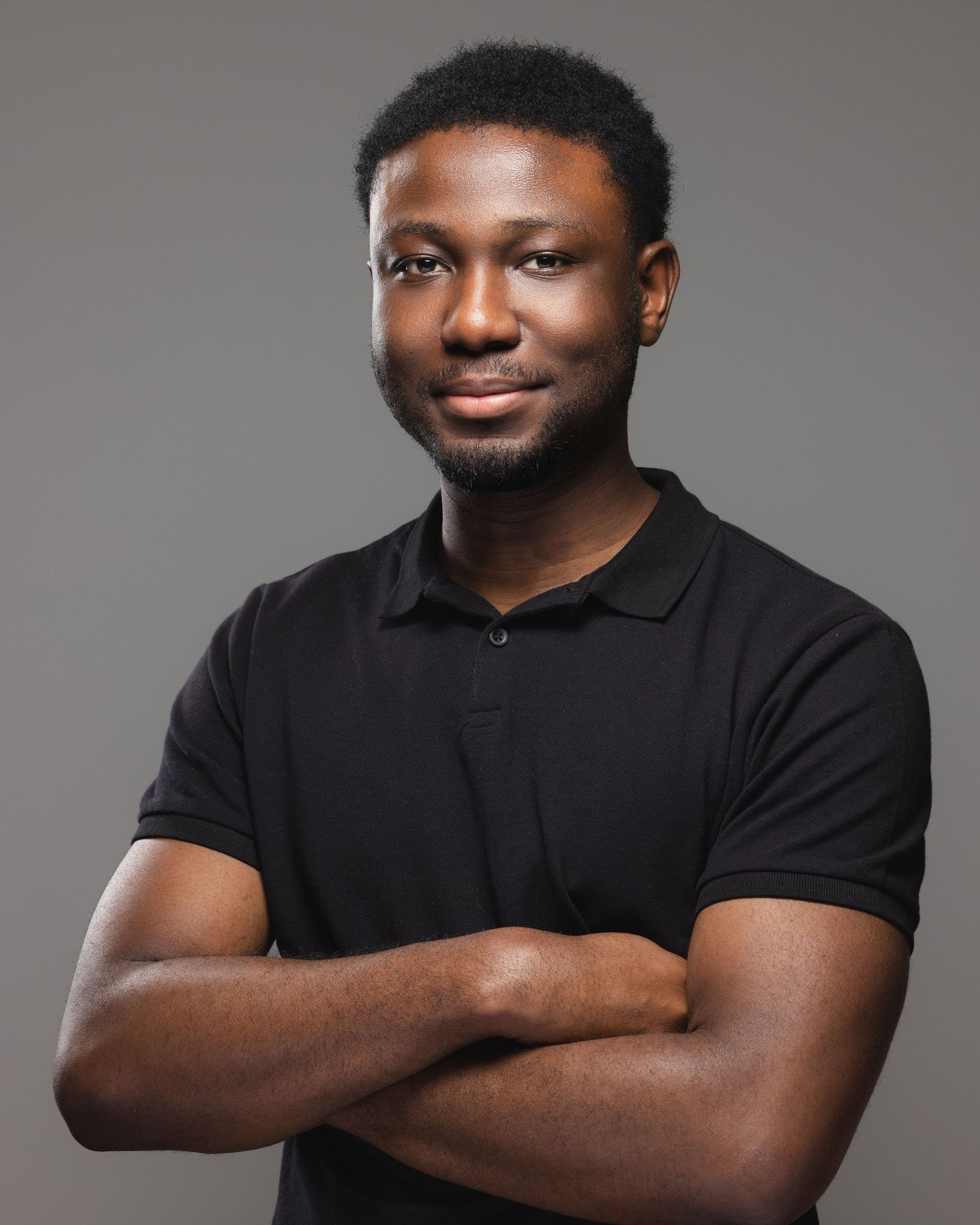 Young Black man in black polo shirt standing with arms crossed, smiling confidently against gray background.