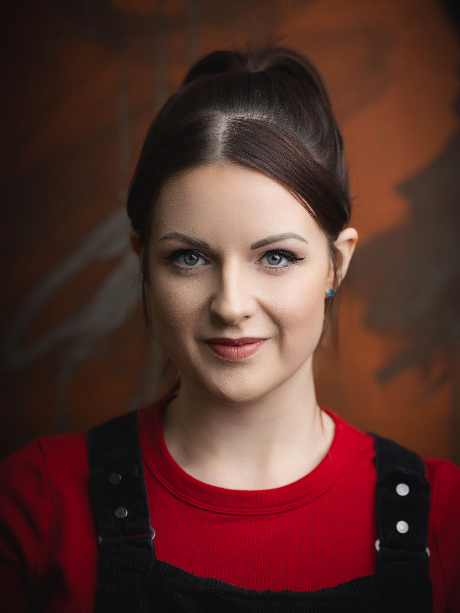 Young woman with dark hair in updo, blue eyes, red top, black overalls, posing against rustic brown background.