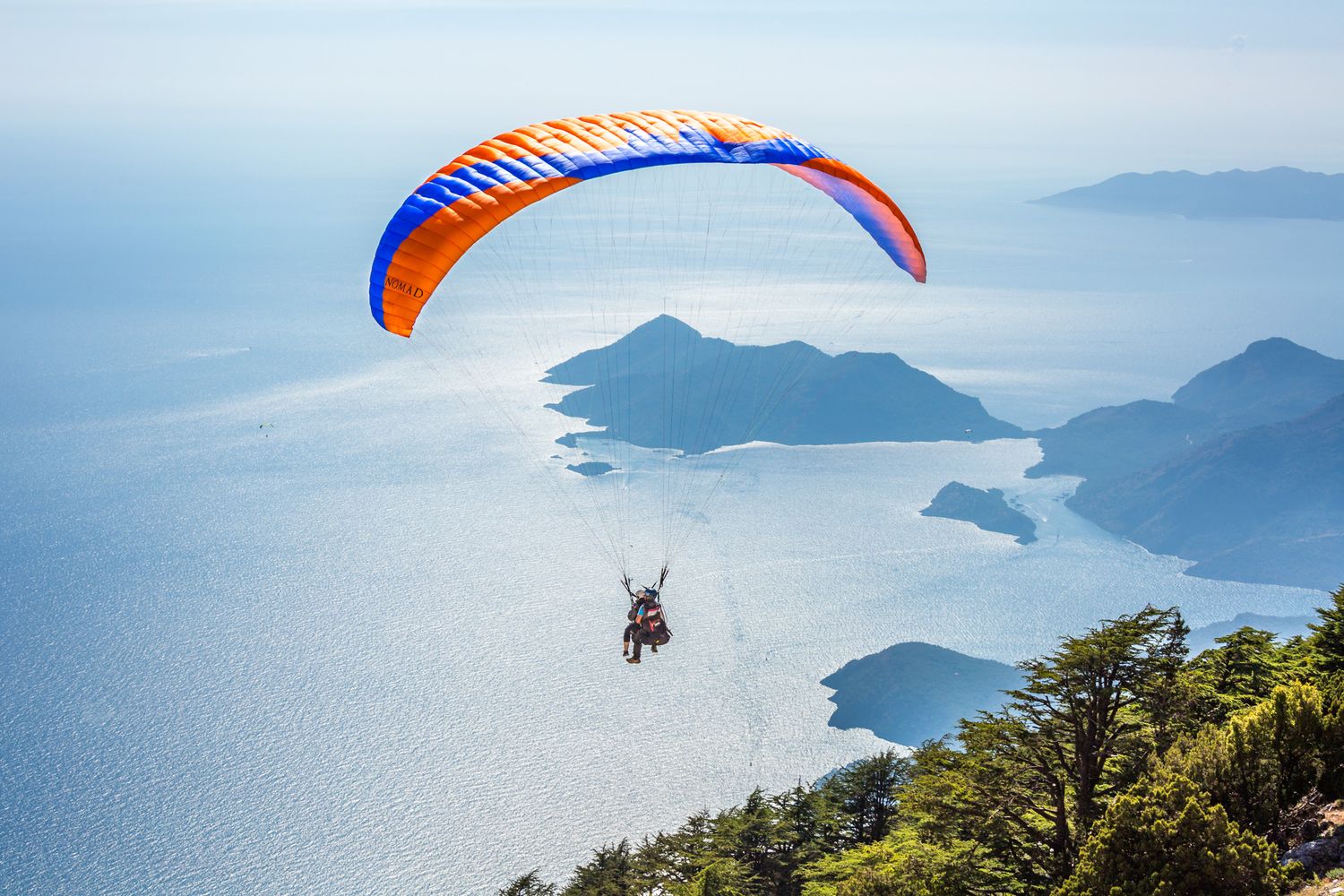 A paraglider soars over a scenic mountain coastline with blue ocean waters and islands visible below.