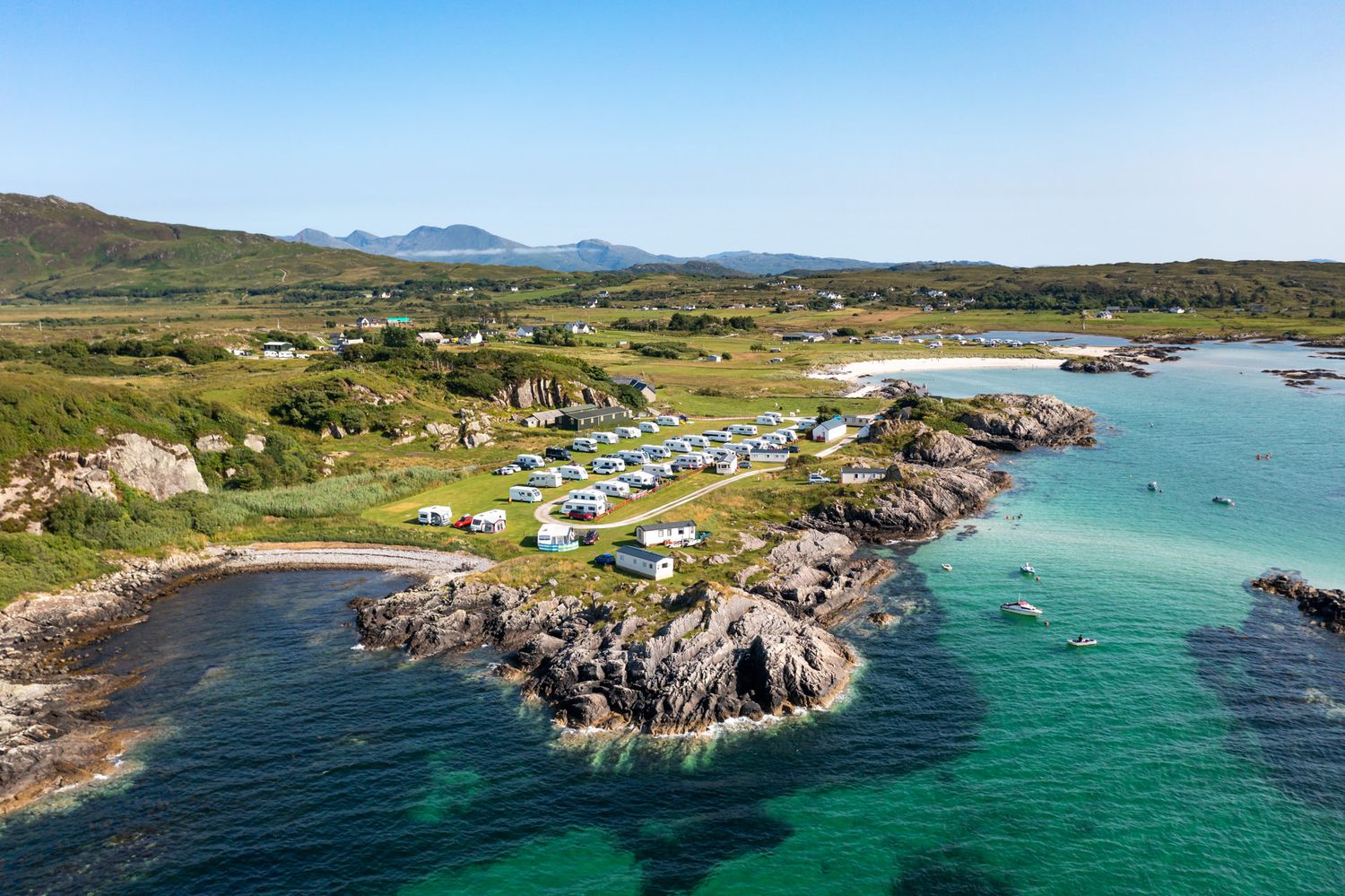 Aerial panoramic view of a campsite with turquoise waters and rocky coastline with camper vans along the Scottish coastline.