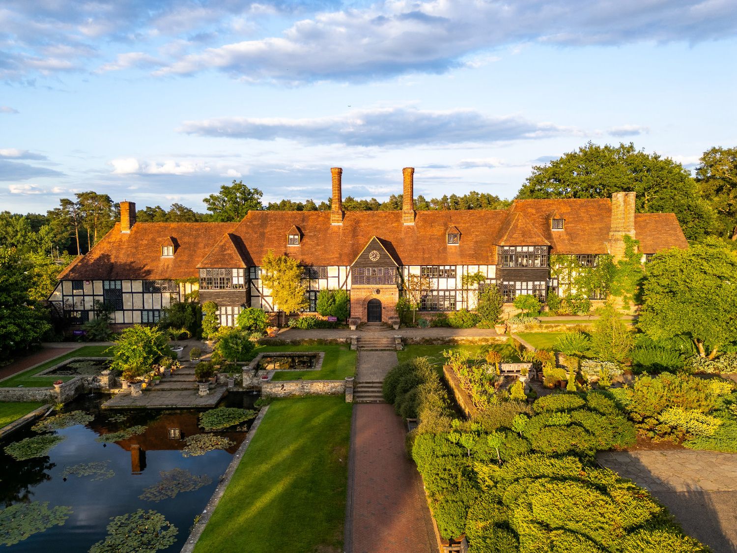 Panoramic view of a traditional Tudor-style estate with manicured gardens and brick chimneys under a cloudy blue sky.
