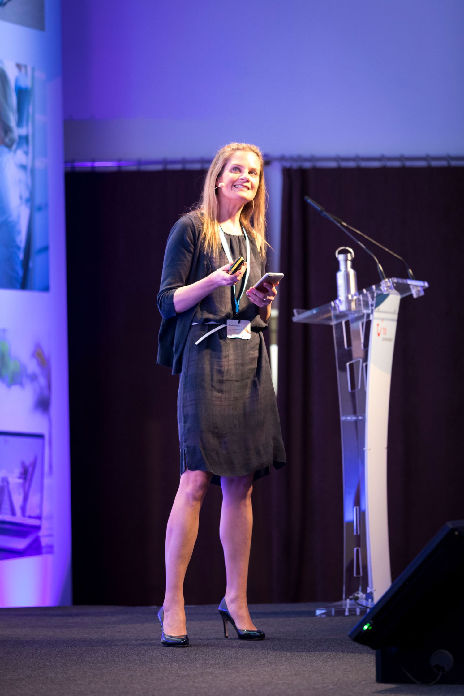 Woman speaker holding a phone presents on stage at a professional conference near a clear podium.