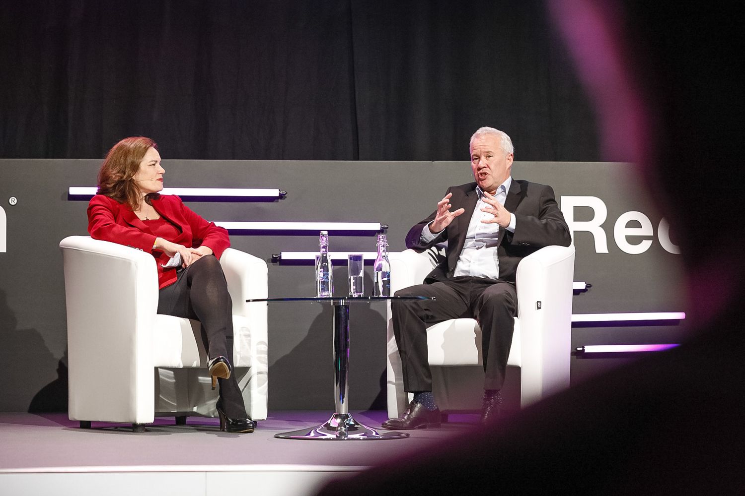 Two professionals engaged in a panel discussion on stage, seated in white chairs at a conference event.