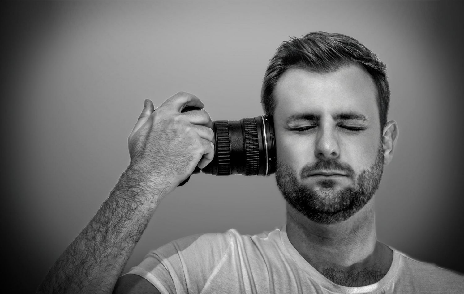 Black and white photo of a man holding a camera lens up to his temple with eyes closed.