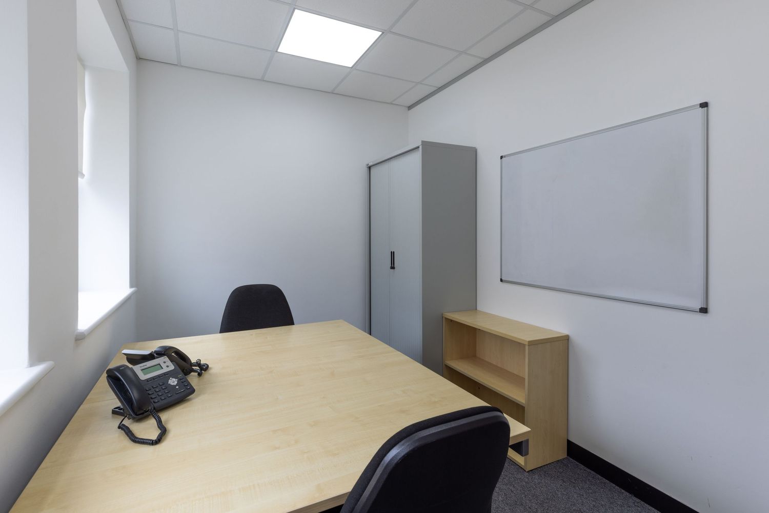 Small office room with a wooden desk, black chairs, whiteboard, storage cabinet, and bookshelf under bright ceiling lights.