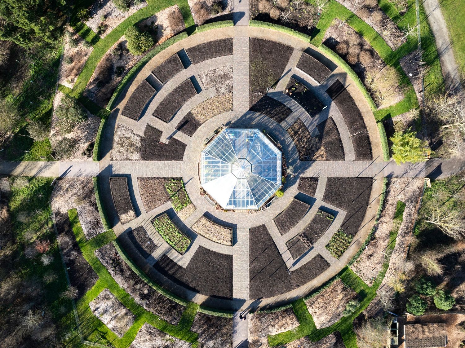 Aerial drone photograph of a formal garden featuring geometric pathways and a glasshouse set within a circular landscaped design.