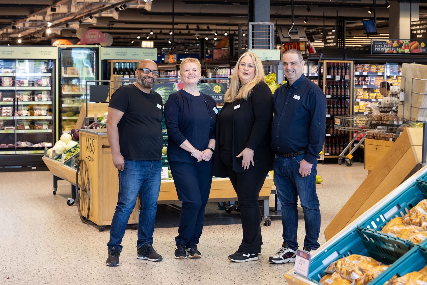 Corporate team photography of four supermarket employees smiling together beside fresh produce and bakery displays.