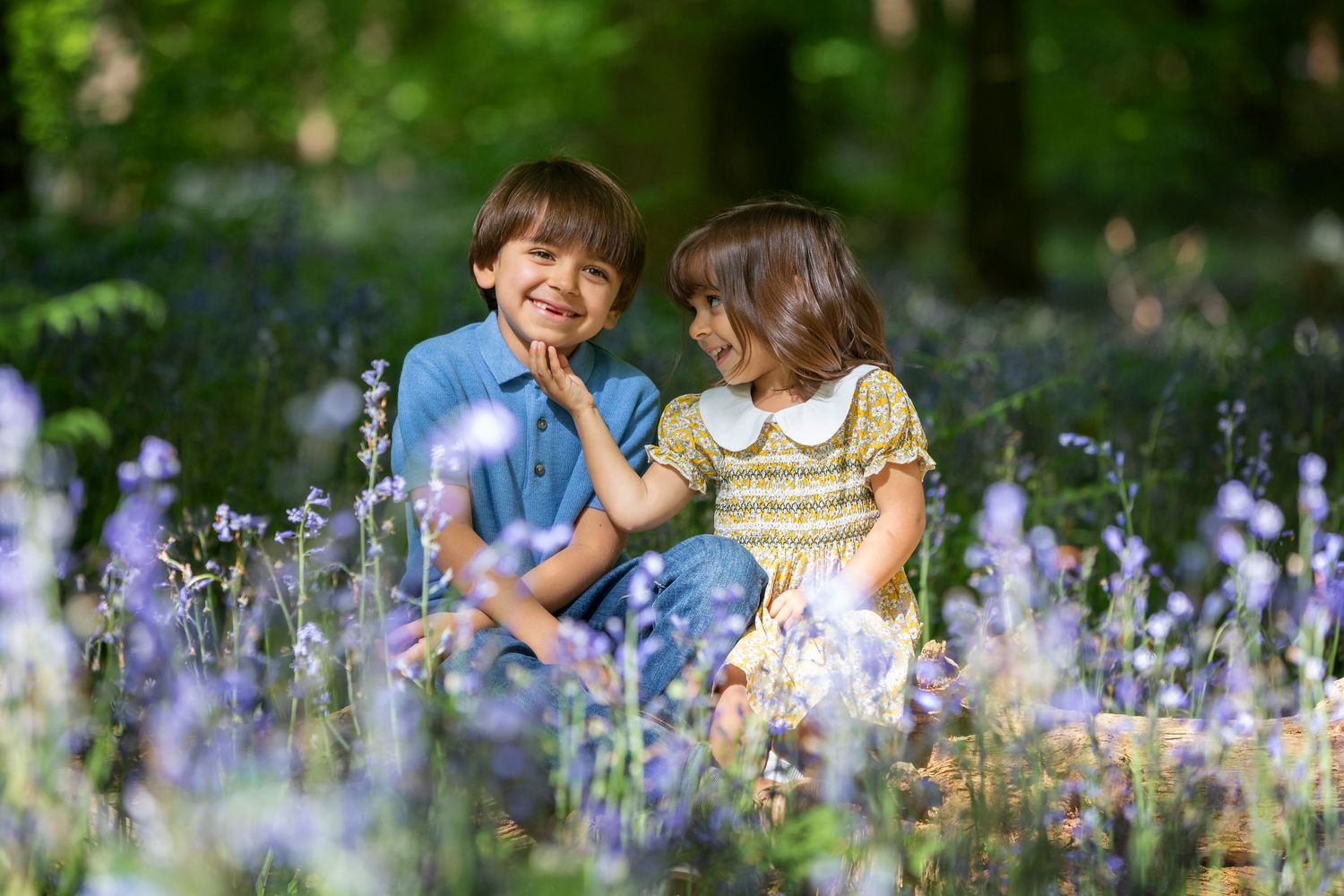 Two happy children sitting together among purple wildflowers in a sunlit forest, smiling and sharing a joyful moment.