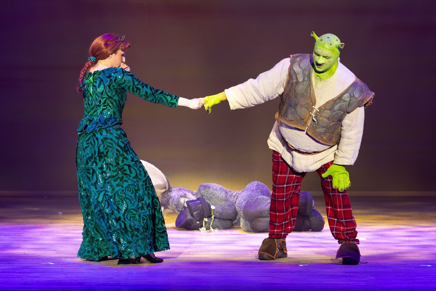 Shrek and Princess Fiona hold hands on stage in a colourful scene during Shrek The Musical at Castle Theatre Wellingborough