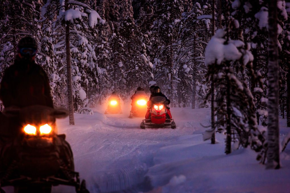 Illuminated snowmobiles travel through a snowy forest path at dusk, creating a magical winter scene in Lapland, Finland.