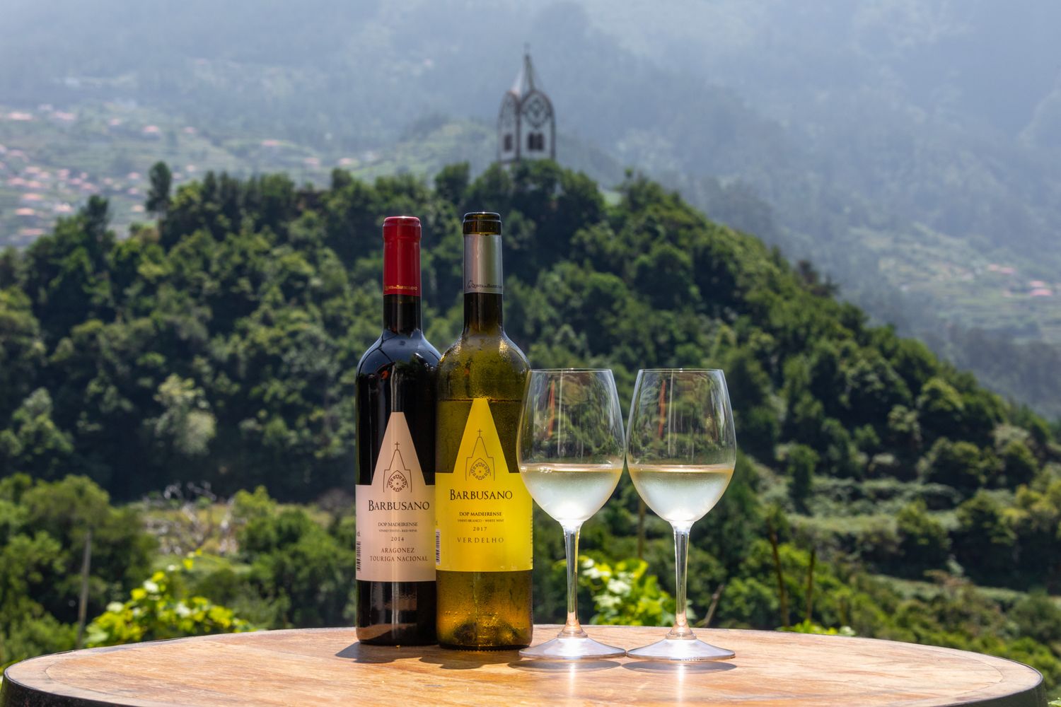 Wine bottles and glasses displayed on table overlooking mountain chapel in scenic landscape.