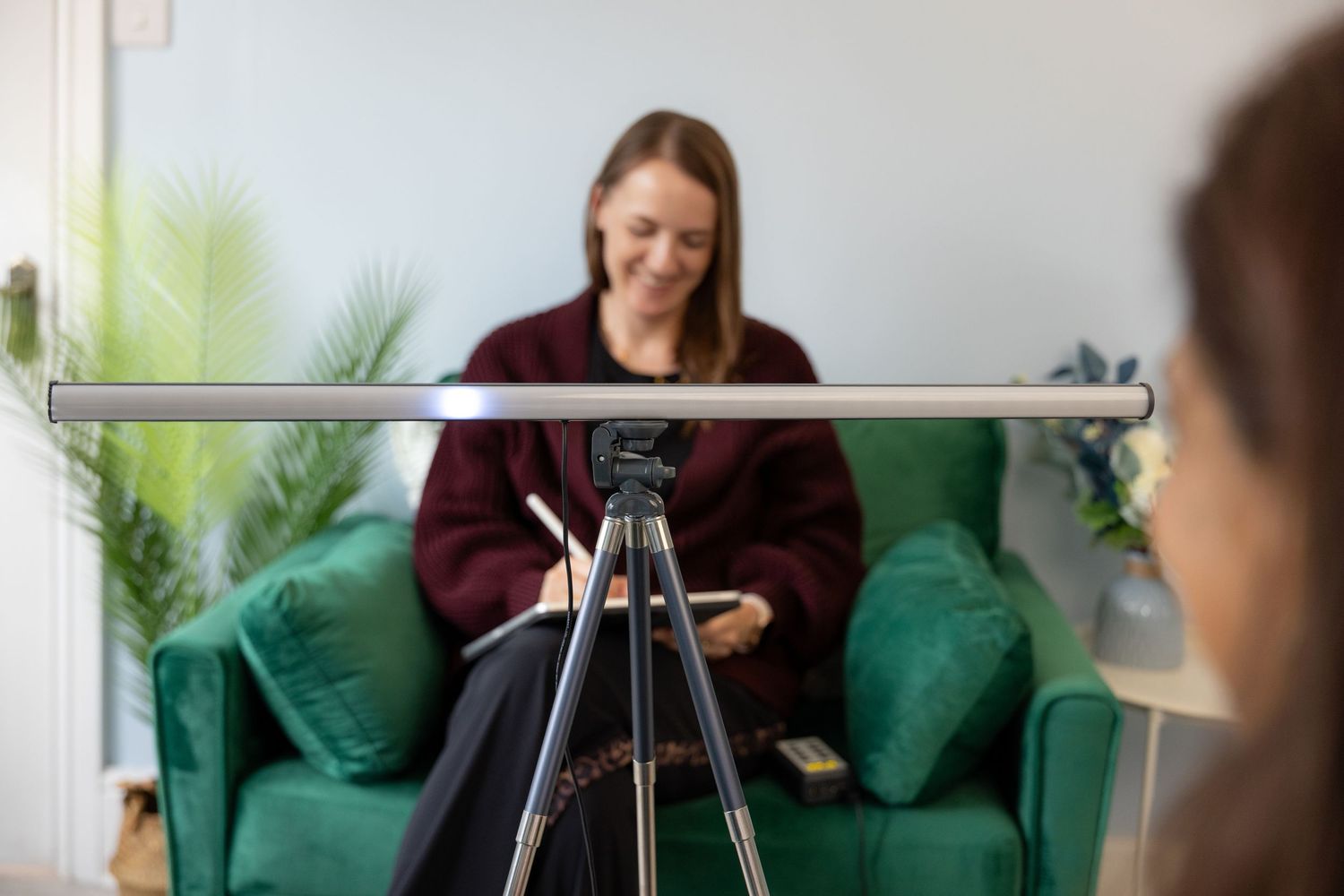 Woman sitting in green armchair smiling, filmed on tripod camera in bright modern room with plants.