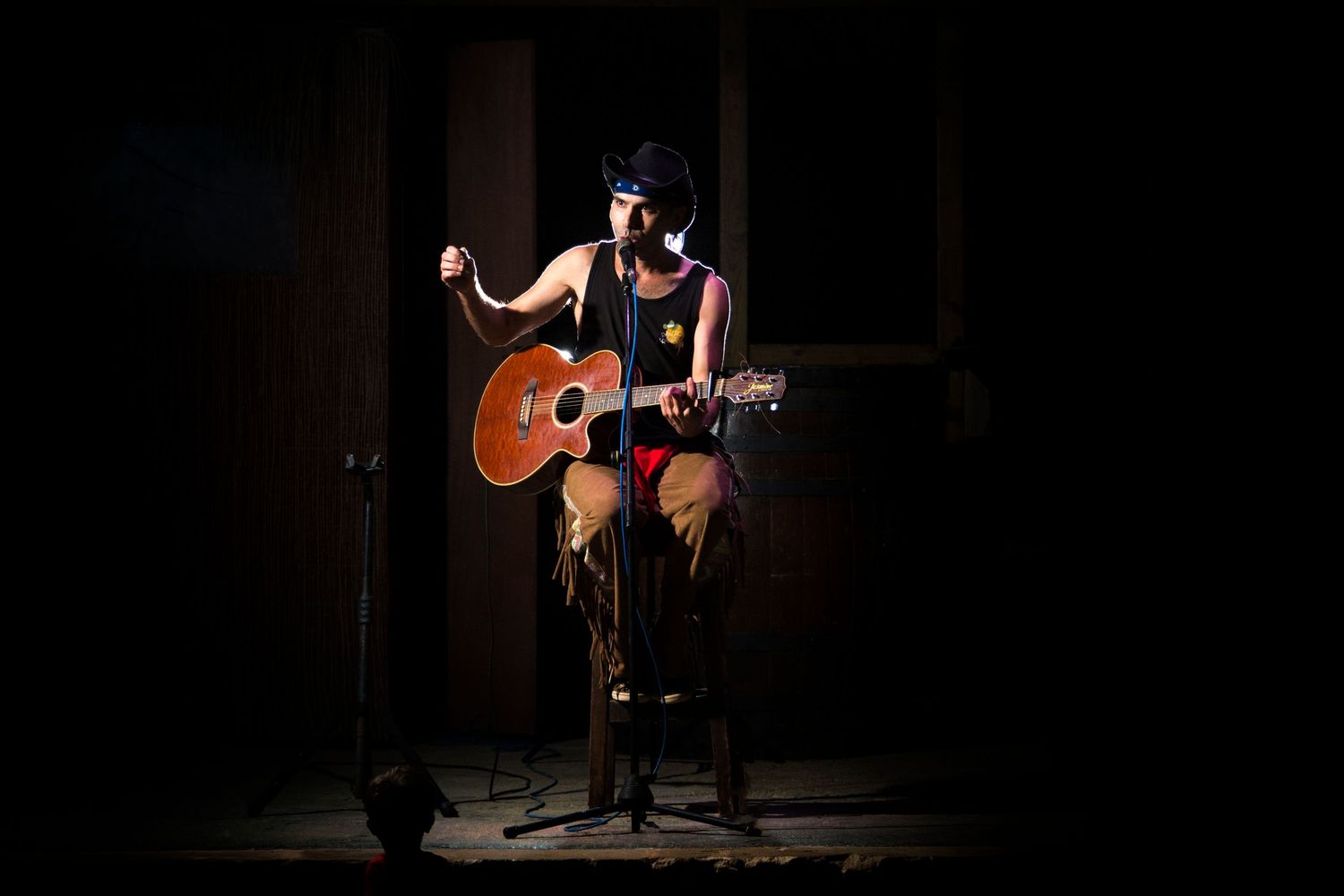Musician sits on a stool playing an acoustic guitar under a dramatic spotlight on a dark stage, singing into a microphone.