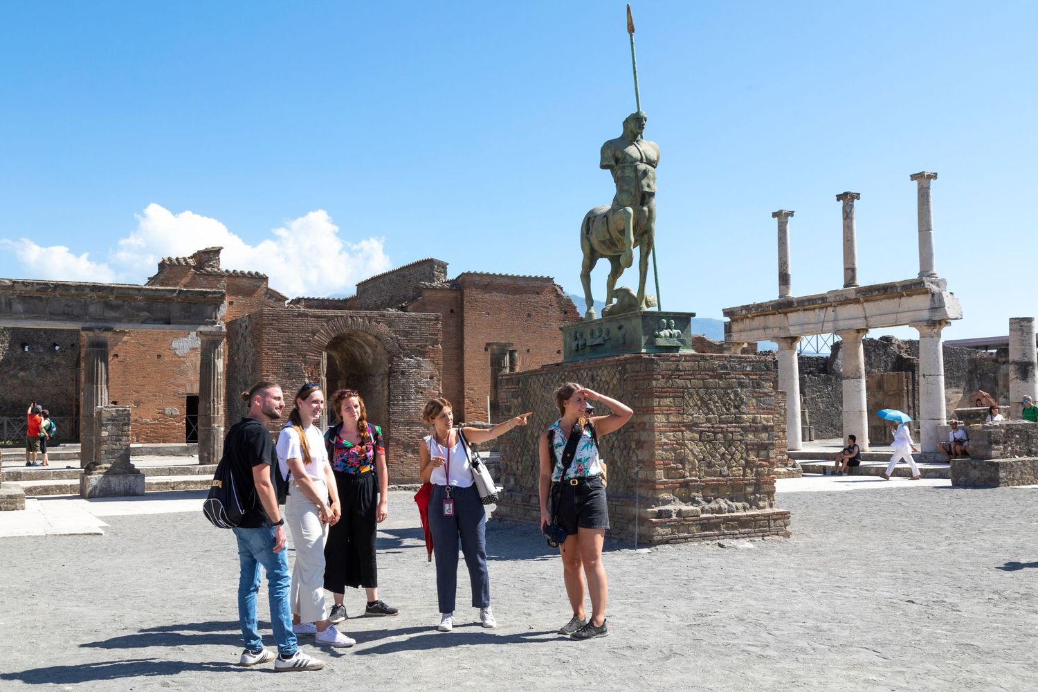 Tourists gather in the ancient Forum of Pompeii, admiring a bronze statue on a stone pedestal beside weathered Roman ruins and columns.