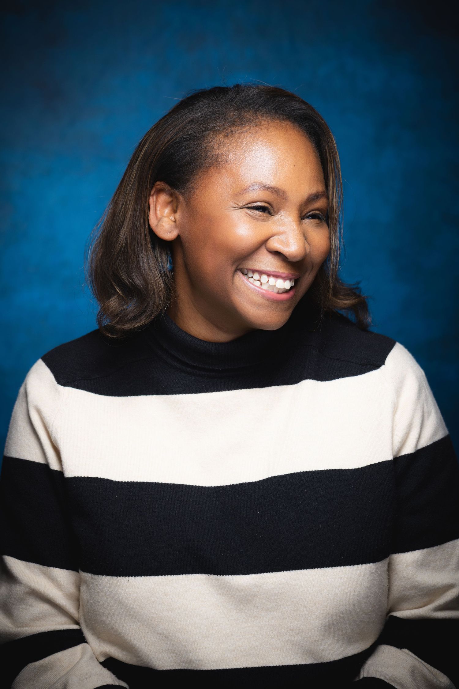 Smiling Black woman in striped black and white turtleneck sweater poses for portrait against blue background.