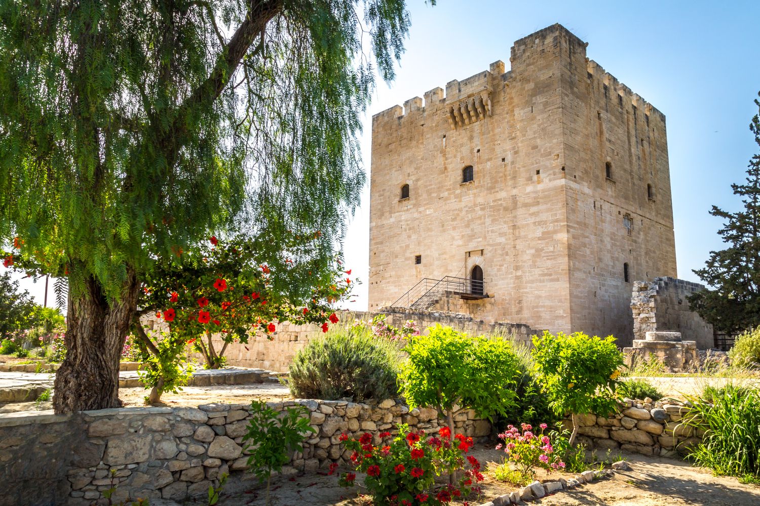 Medieval stone tower with colorful flowers and gardens at Kolossi Castle in Cyprus on sunny day.