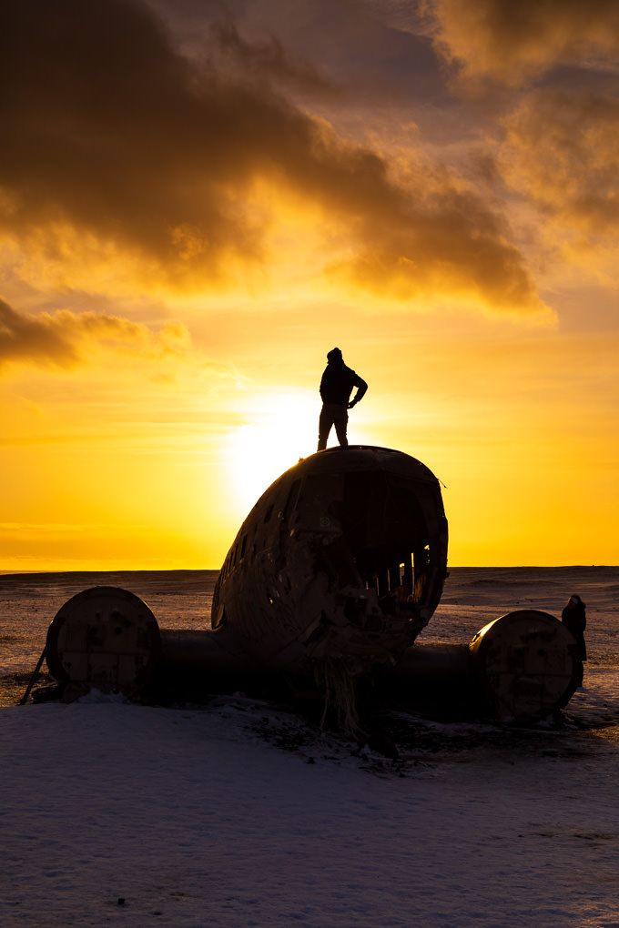 Travel photography of a silhouetted figure stands atop Iceland's Sólheimasandur Plane Wreck at golden hour.