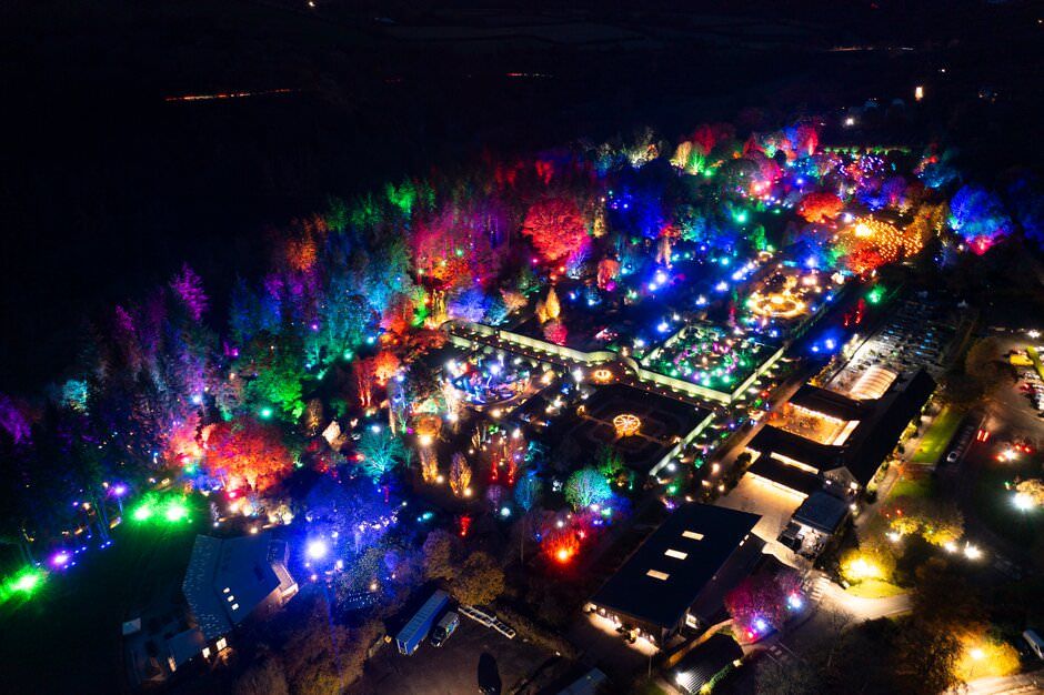 Aerial view of a vibrant nighttime festival with colorful lights illuminating trees and vendor booths in a dark landscape.