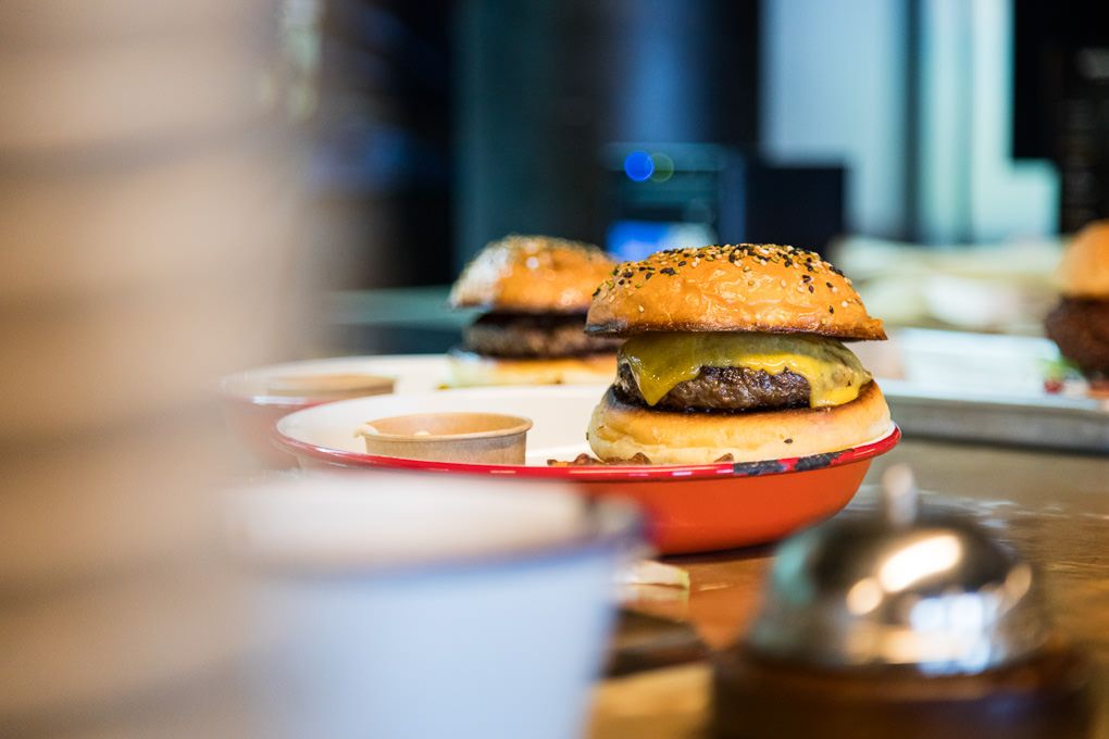 Two juicy cheeseburgers with sesame seed buns on orange plates sit on a wooden counter in soft focus. Le Kok restaurant, Iceland.