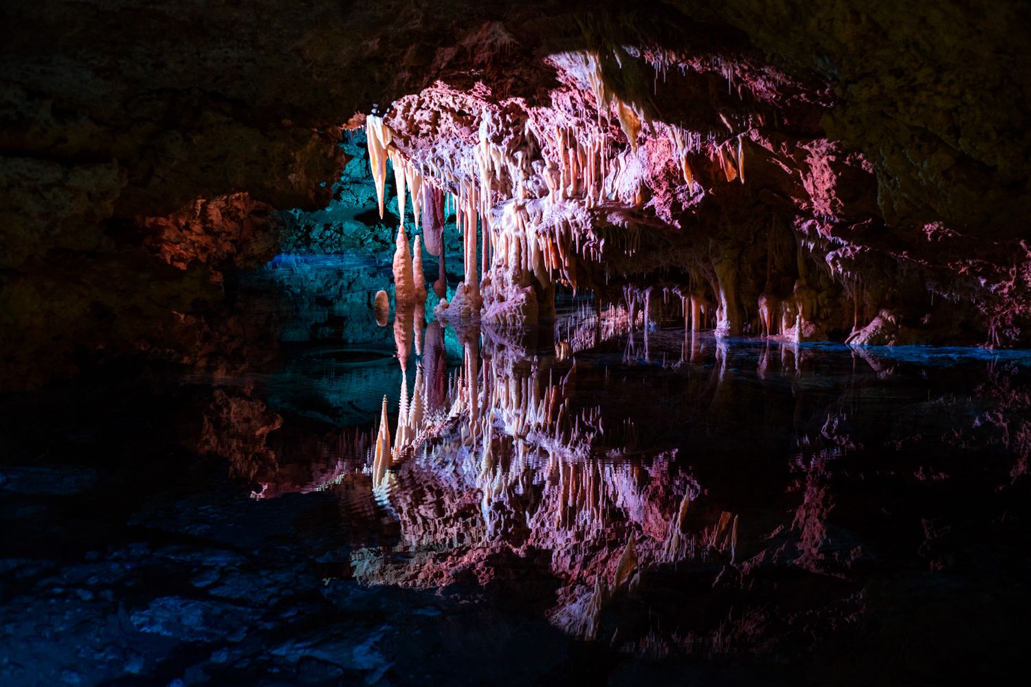 Dramatic stalactites reflecting in dark cave pool with pink and blue illumination create a surreal mirror effect.