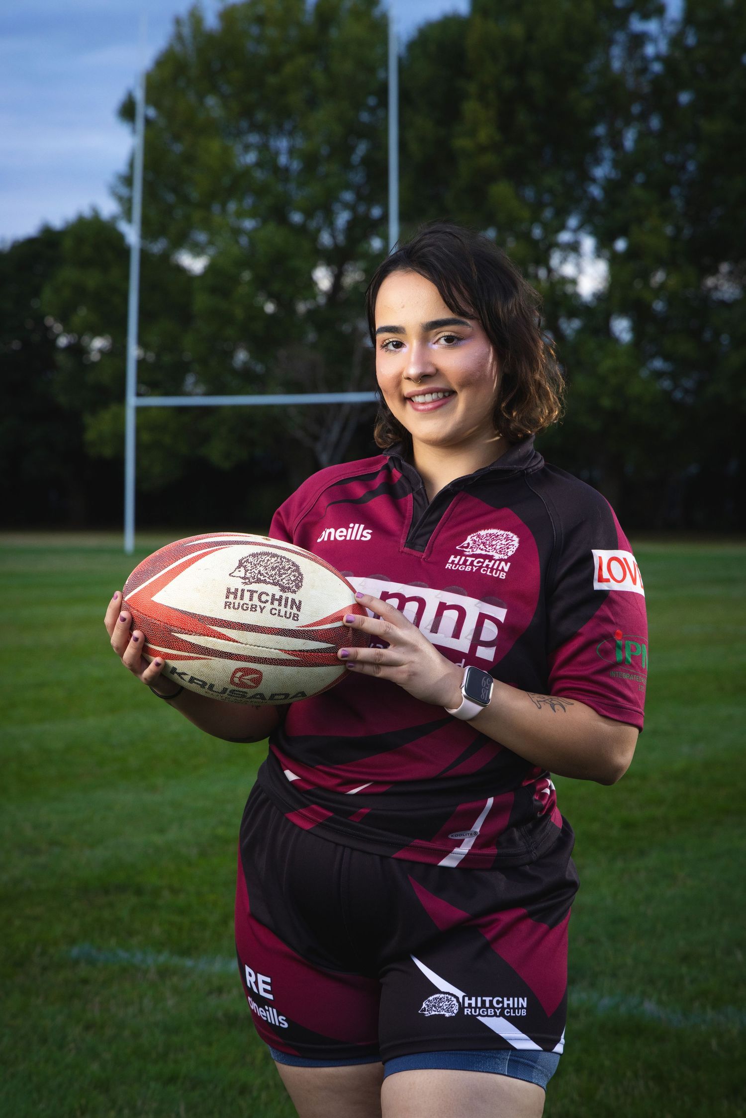 Young woman in Hitchin Rugby Club kit holding a rugby ball on a pitch with goalposts in background.
