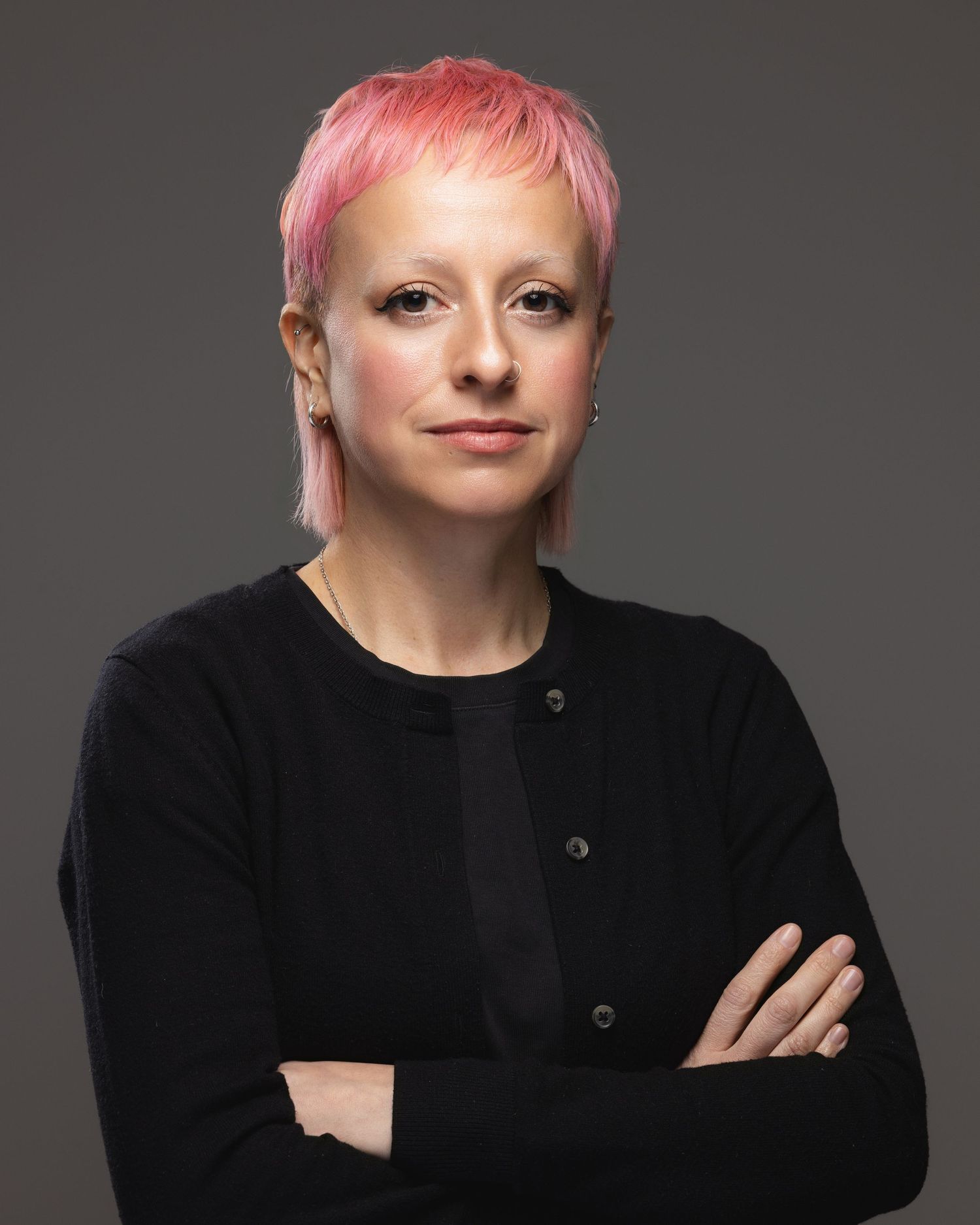 Woman with pink pixie cut hair, wearing black cardigan, arms crossed, professional headshot on gray background.