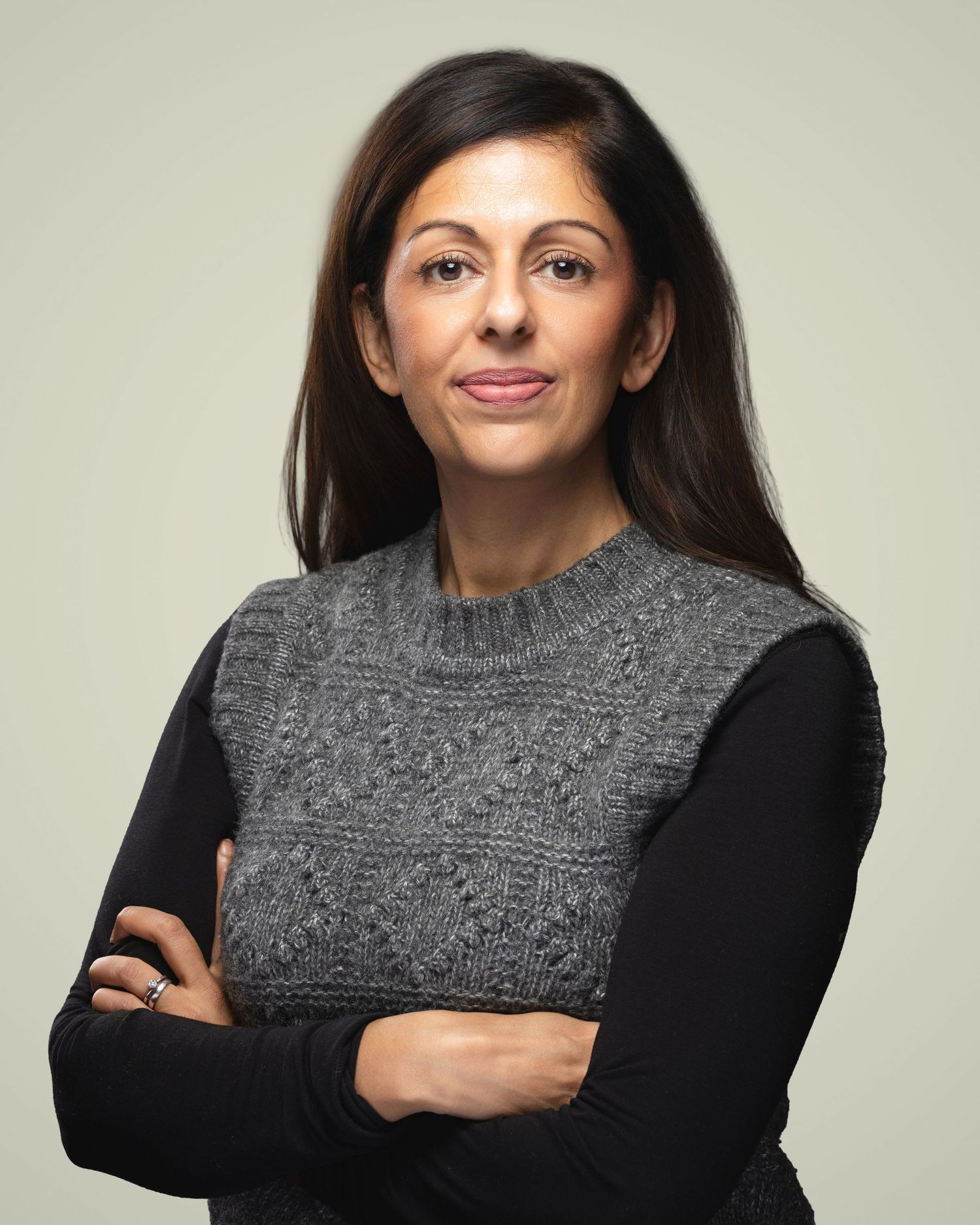 Woman with dark hair poses confidently with arms crossed, wearing a gray knit vest over black long-sleeve top.