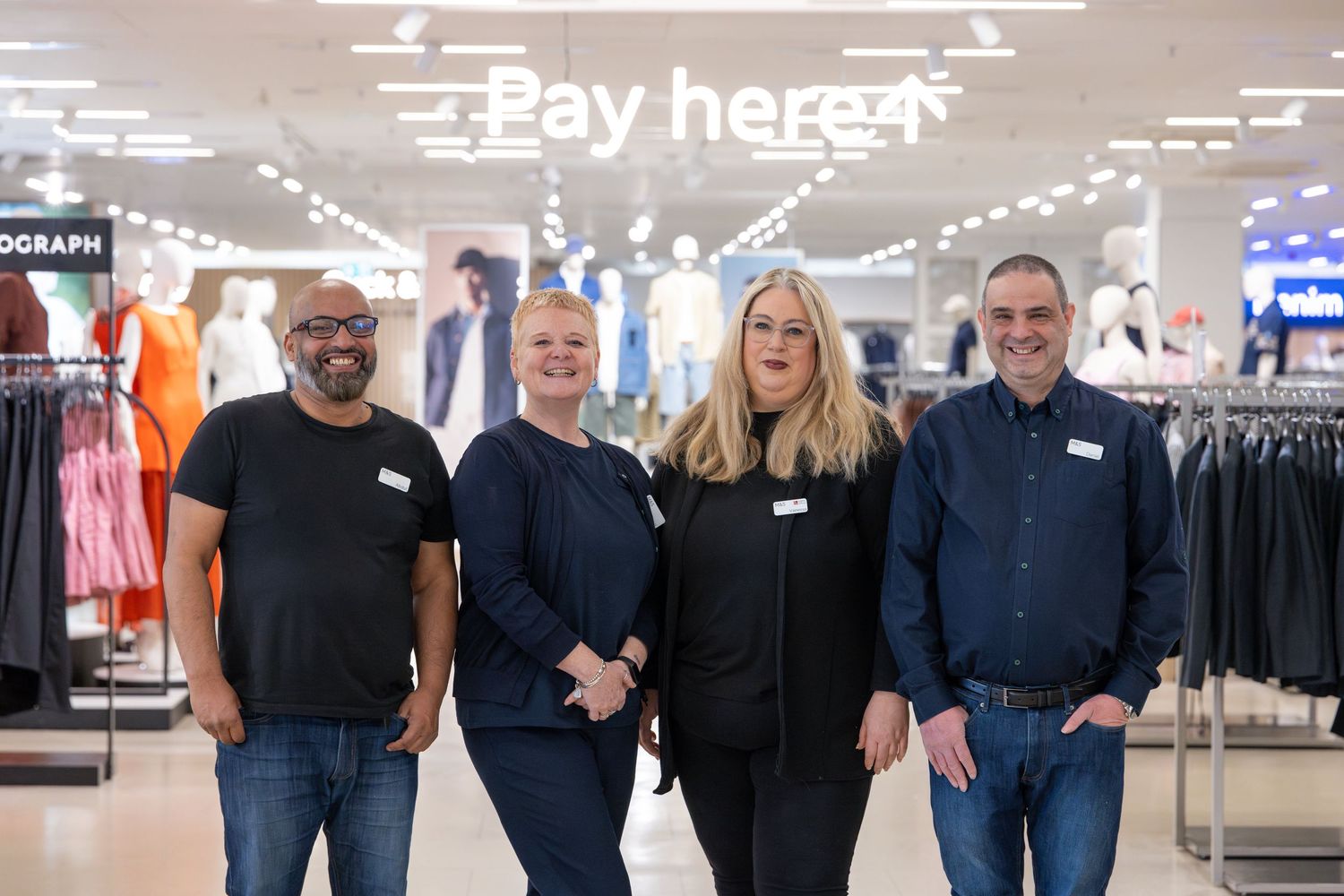 Four retail store employees smiling together in a modern clothing store with a Pay Here sign in the background.