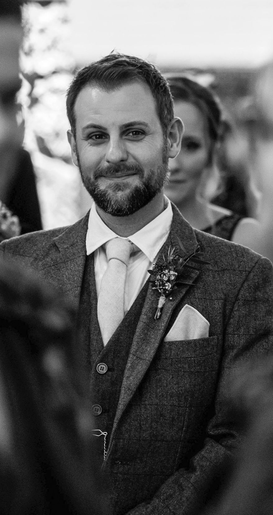 Black and white photo of a bearded groom smiling in a tweed suit with boutonniere at an outdoor wedding.