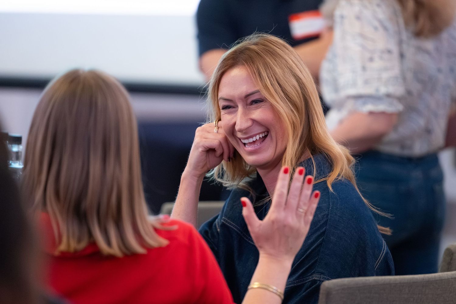 A smiling blonde woman in a navy top laughs during a casual indoor meeting or networking event.
