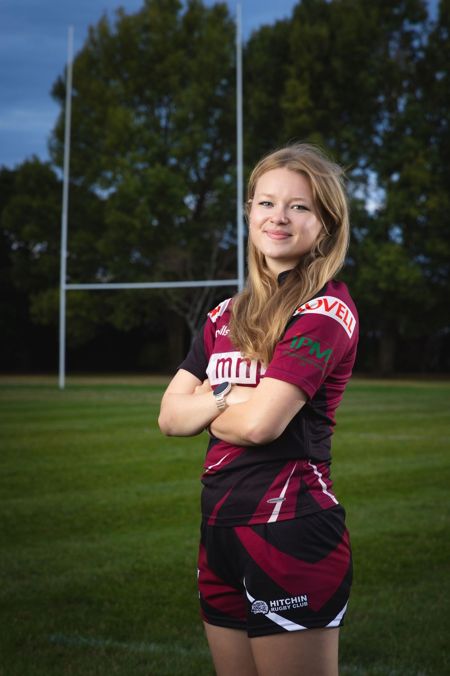 Young female rugby player in Hitchin Rugby Club jersey posing with arms crossed in front of goalposts on a field.