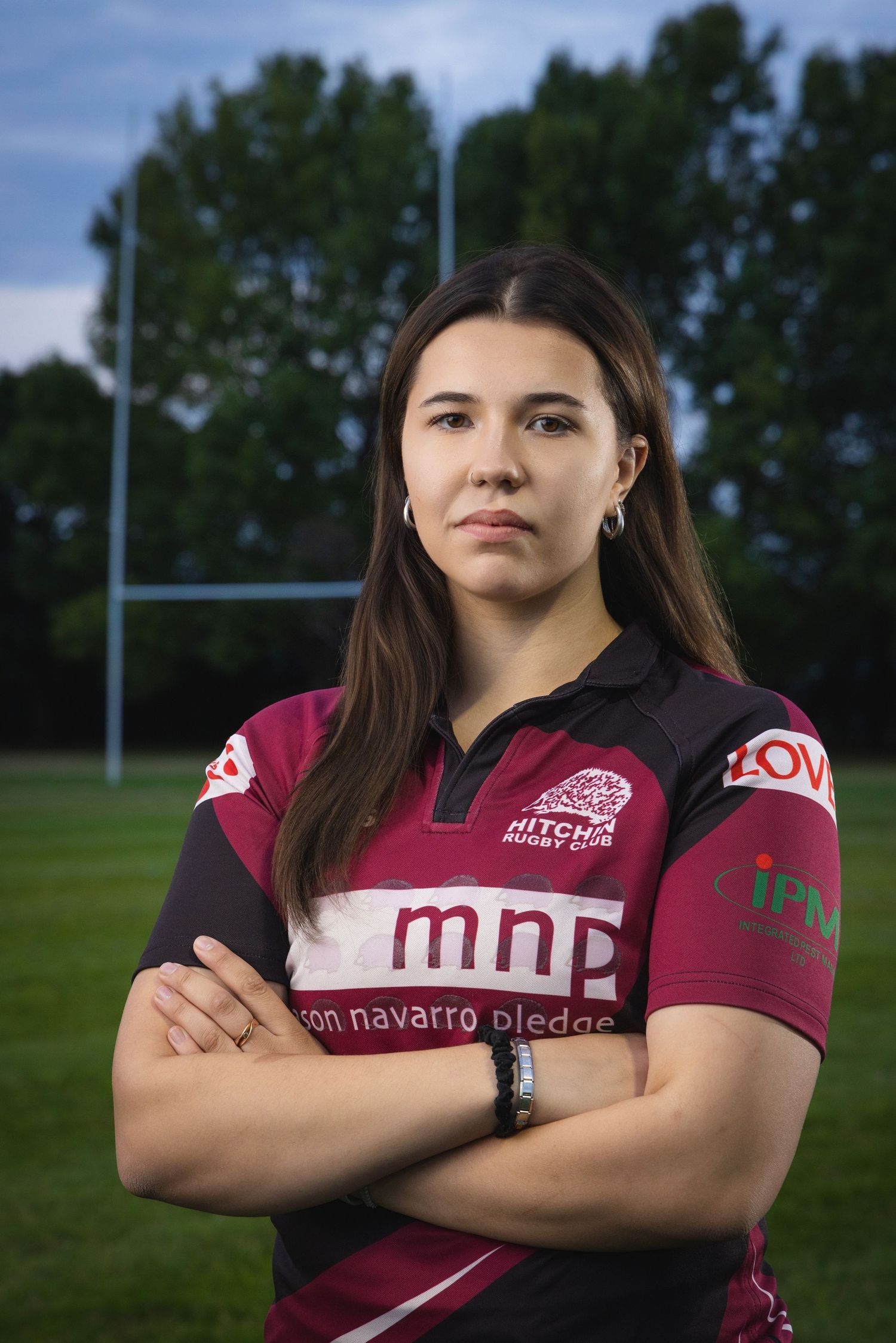 Young woman in Hitchin Rugby Club jersey stands confidently with arms crossed on a rugby field.