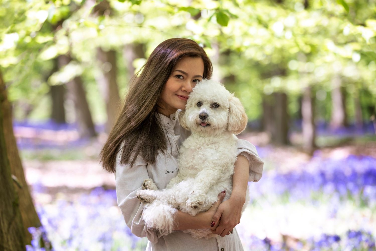 Young woman holding a fluffy white poodle in a bluebell forest with tall green trees in the background.