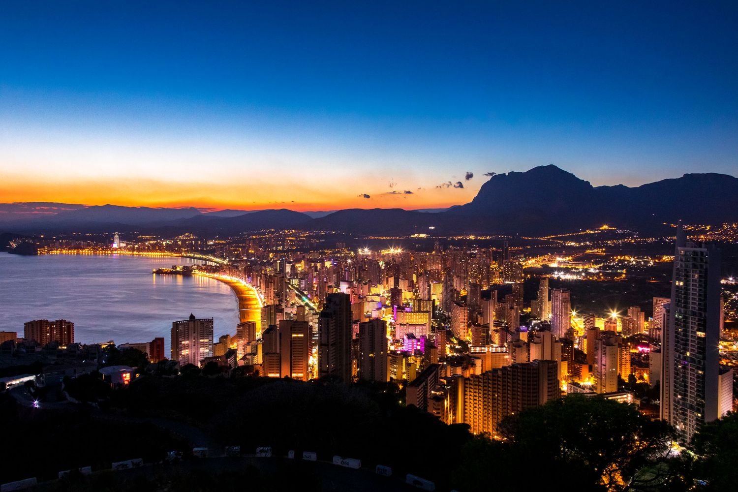 Panoramic night view of illuminated coastal city skyline with mountains and bay at twilight.