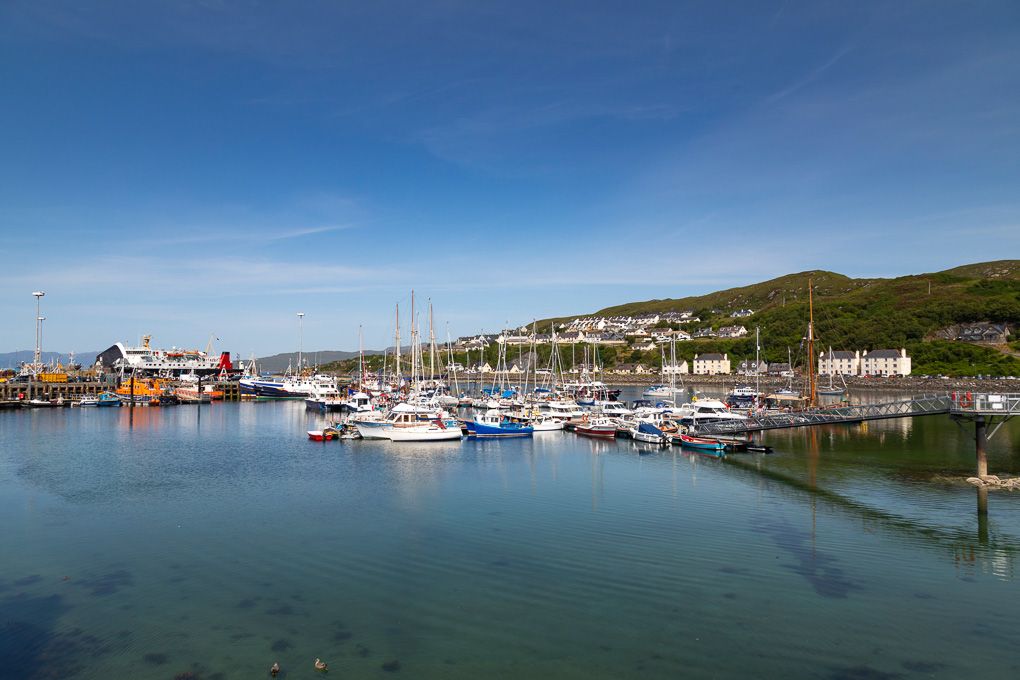 Panoramic view of Mallaig harbour with boats moored in calm blue waters against a scenic coastal landscape.
